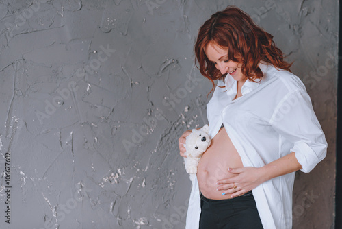 Pregnant woman in a white man's shirt, a studio portrait. Looking at the midsection. With a Teddy bear in front of him.