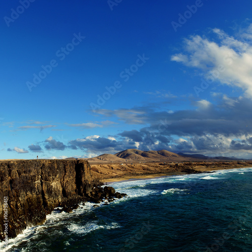 View on a coastline with huge rocks and sandy beach on a sunny evening with soft light. Mountains at horizon and clouds at the sky
