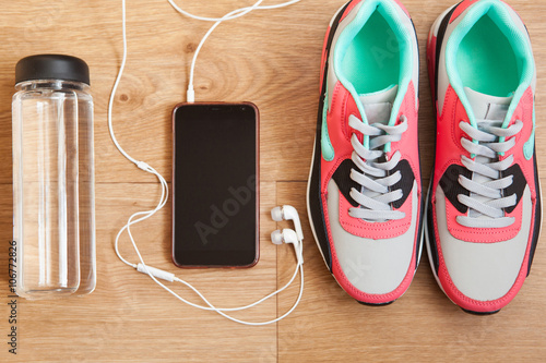 red and grey sneakers with grey shoelaces and bottle with water, mobile phone with white headphones on wooden background indoors