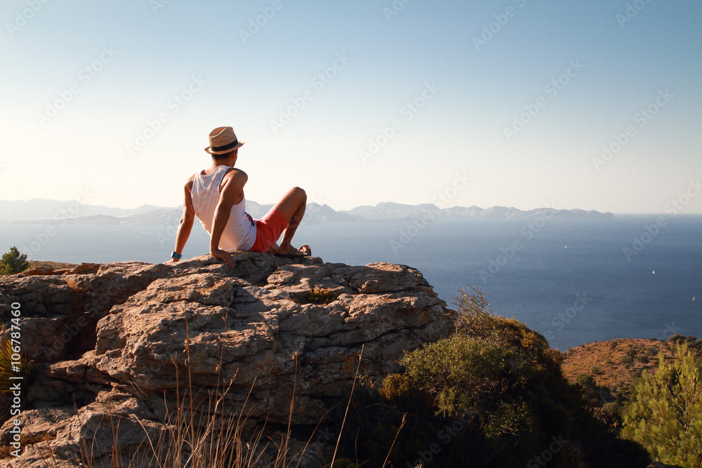 Contemplative young man sitting on the rocks on the top of the mountain at sunset. Looking at distance with the blue sea in front. Wearing a straw hat in summer.