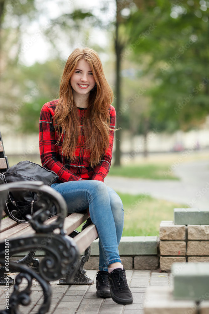 Young smiling redhead woman in red plaid jacket and blue jeans sitting on  park bench with blurred park background Stock Photo | Adobe Stock
