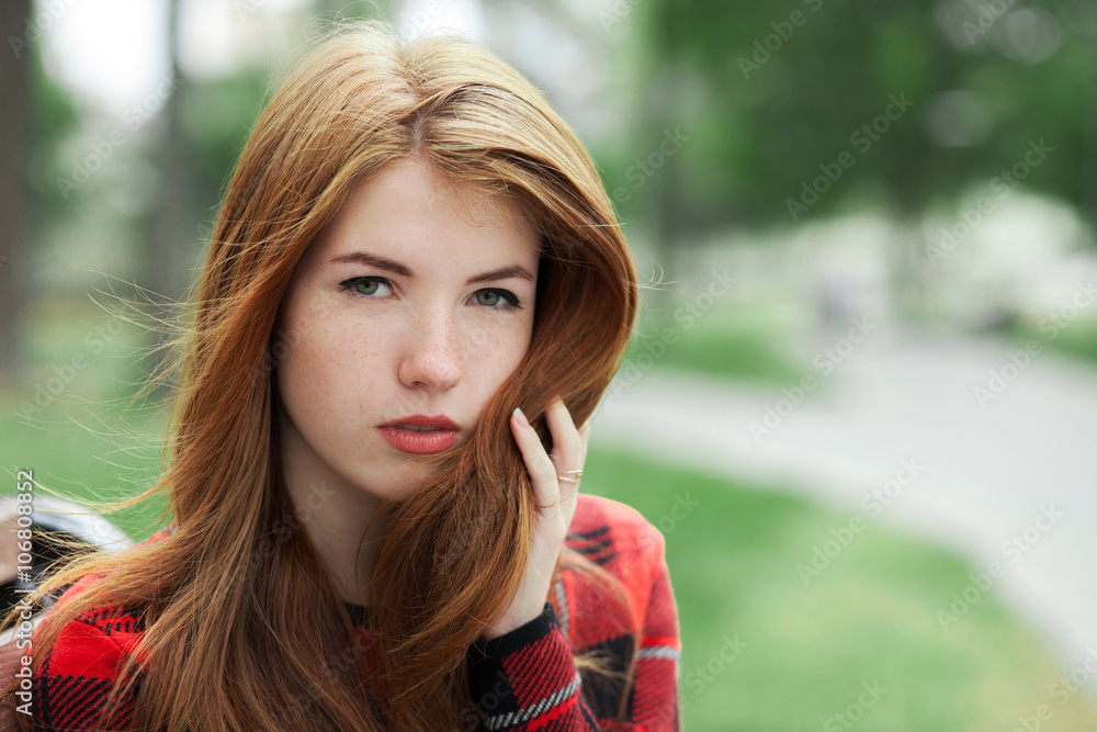 Closeup portrait of young alarmed redhead woman in red plaid jacket touching her hair with blurred park background Closeup portrait of young alarmed redhead woman in red plaid jacket touching her hair with blurred park background