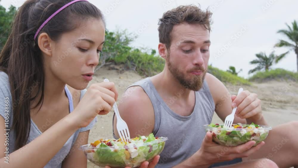 Salad - healthy fitness woman and man couple talking and eating food lunch sitting on beach after workout. Mixed race Asian Caucasian female model and male models in sportswear. Salad - healthy fitness woman and man couple talking and eating food lunch sitting on beach after workout. Mixed race Asian Caucasian female model and male models in sportswear.