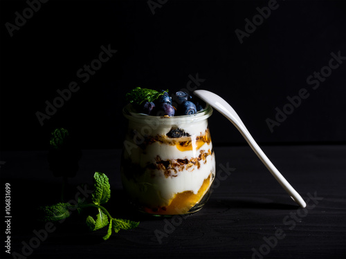 Yogurt oat granola with jam, blueberries and mint leavesin glass jar on black backdrop photo