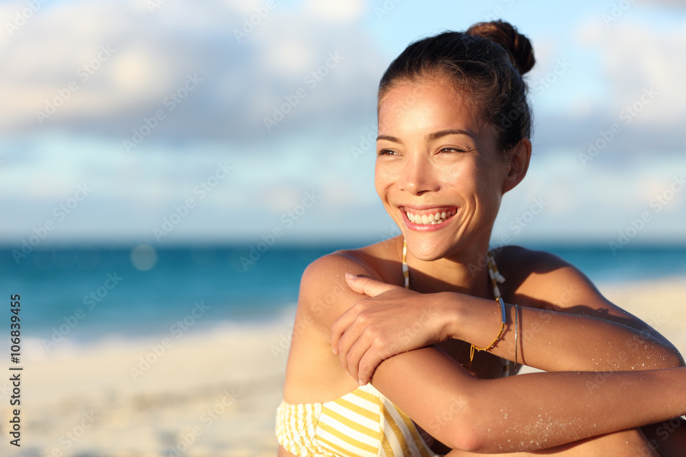 Happy healthy young Asian chinese woman smiling on beach enjoying relax lifestyle sitting in swimwear. Beautiful mixed race tourist in hair bun and golden jewelry at sunset on Caribbean travel.