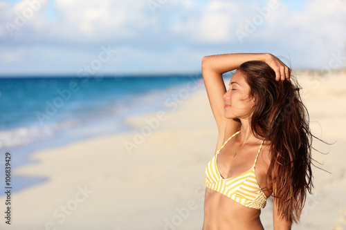 Carefree bikini woman enjoying sunset on beach. Beautiful relaxing Asian model caressing hair with closed eyes over the ocean on summer travel vacation in yellow swimsuit top in tropical destination.