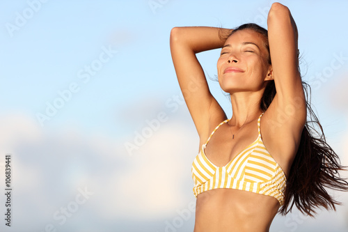 Relaxing bikini woman feeling good and free in outdoor nature breathing fresh air with arms up showing healthy body and smooth skin and armpits for laser epilation treatment. Asian model on beach. photo