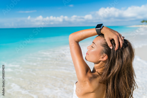 Smartwatch woman relaxing on beach wearing smart wrist watch for activity tracker for an active lifestyle on tropical Caribbean travel destination during summer vacation. Sexy Asian model sun tanning.