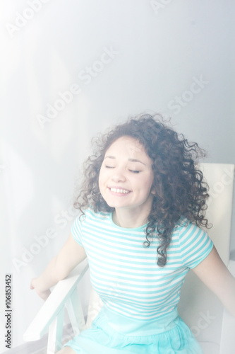 Portrait of a happy young girl with curly hair in a white chair. Morning room and light. Misty picture. Enjoy life