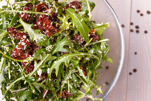 Green healthy salad with arugula and sun-dried tomatoes and parmesan cheese on a wooden kitchen table. Pepper and seasonings.  Italian cuisine. Top view.