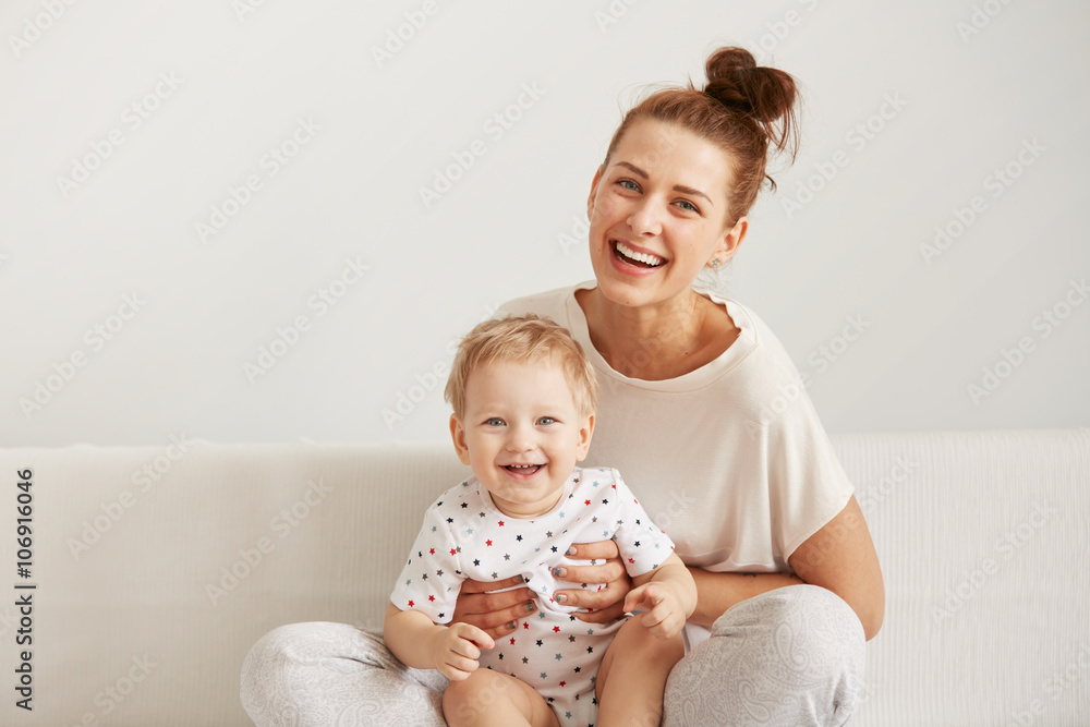 Mother and baby laugh together at home. They are sitting on the sofe in a brightly lit living room at the weekend together, lazy morning, warm and cozy scene. Selective focus Mother and baby laugh together at home. They are sitting on the sofe in a brightly lit living room at the weekend together, lazy morning, warm and cozy scene. Selective focus