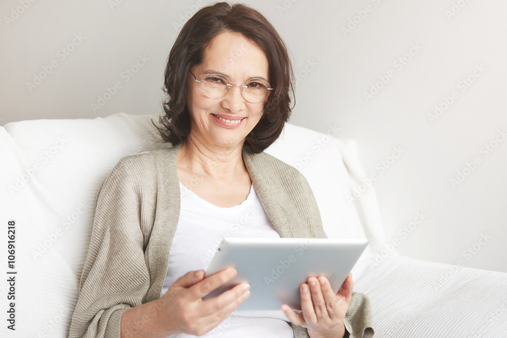 Middle-aged woman using touch pad reading a message, email or information on her tablet computer sits on a couch at home. Smiling senior woman at home connected on internet. Middle-aged woman using touch pad reading a message, email or information on her tablet computer sits on a couch at home. Smiling senior woman at home connected on internet.