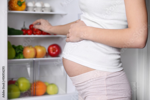 Pregnant woman standing near refrigerator full of healthy food