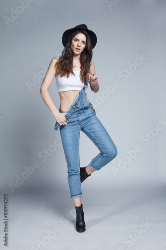 Beautiful young woman showing her emotion in studio shot. Model wearing nice blue suspender jeans, white t-shirt and black hat. Outfit is complete with rings, necklaces and gorgeous smile