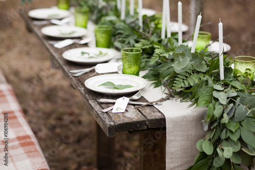 Weddind table setting with white plates and green glasses decorated with white candles, green leaves and eucalyptus and linen tablecloth outdoors
