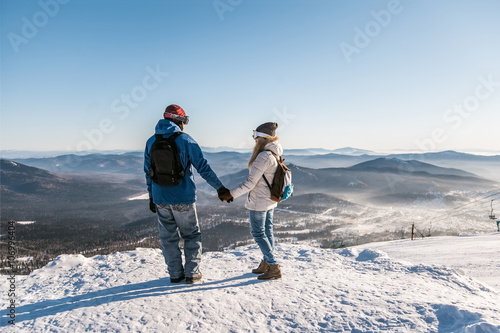 Girl and guy with backpacks in masks for snowboarders holding hands on the background of the ski slopes. 