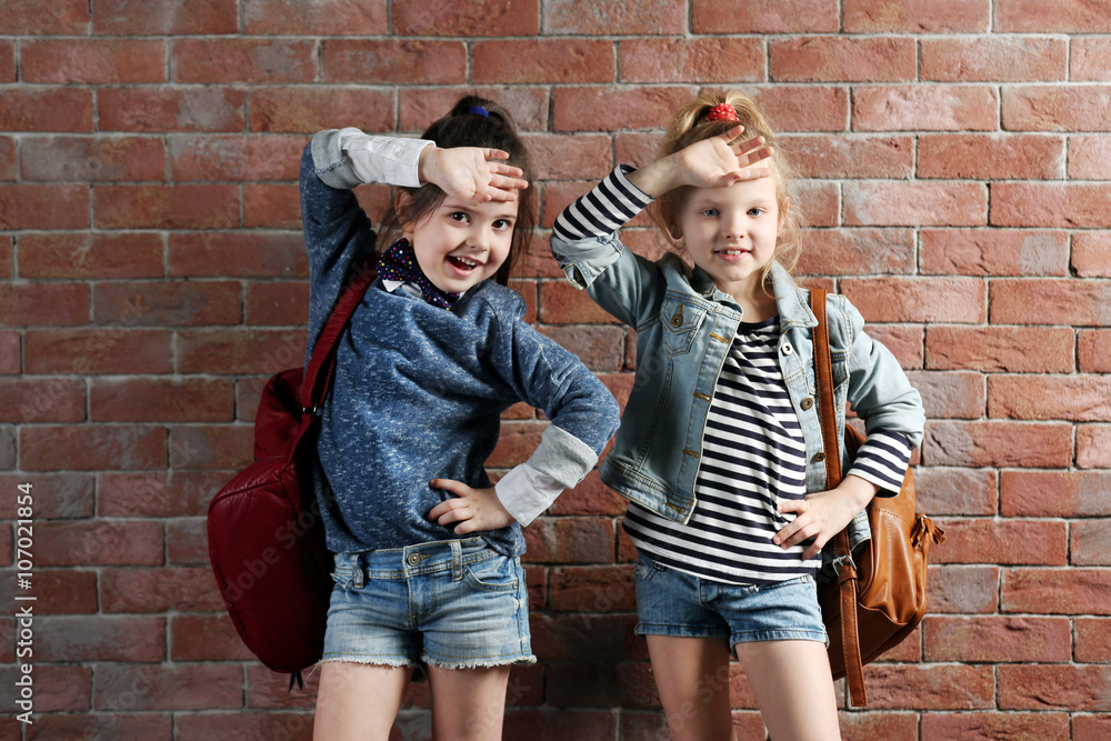 Two stylish little girls with backpacks on wall background