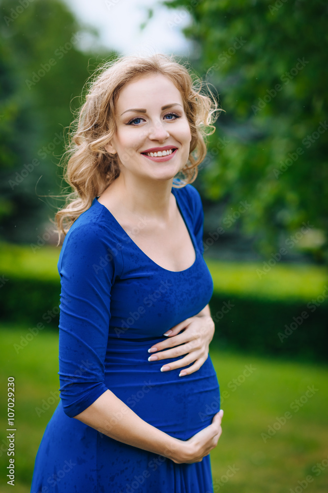 Portrait of beautiful young pregnant woman in blue dress with long blond curly hair holding her belly, smiling and looking at camera in summer park. Pregnancy and femininity concept. Mother's Day Portrait of beautiful young pregnant woman in blue dress with long blond curly hair holding her belly, smiling and looking at camera in summer park. Pregnancy and femininity concept. Mother's Day
