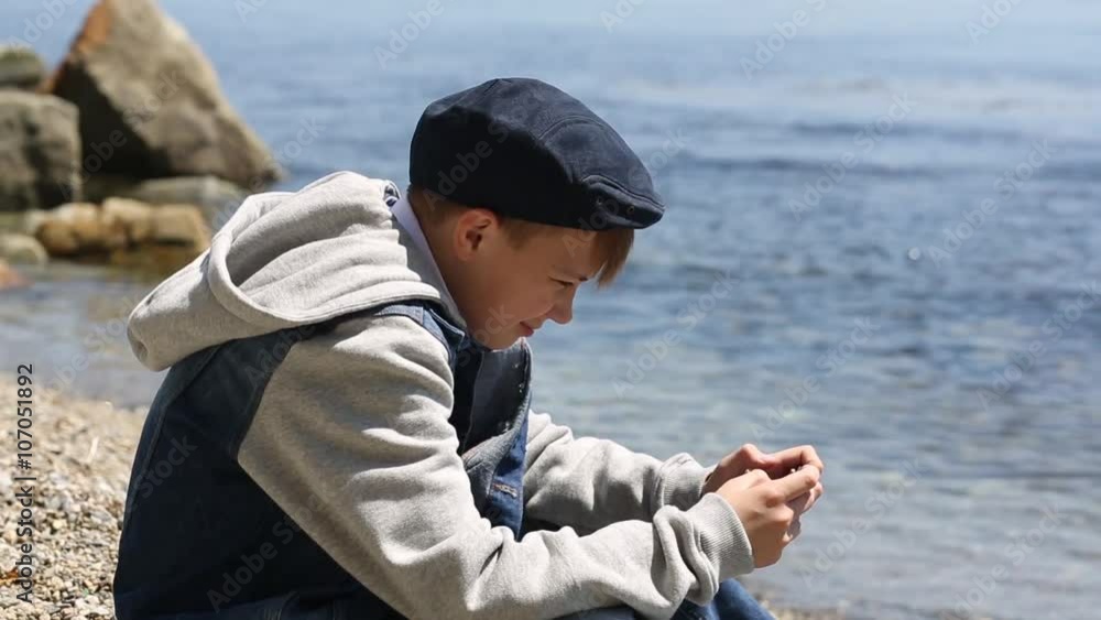 Boy uses a smartphone while sitting on the beach in spring day Boy uses a smartphone while sitting on the beach in spring day