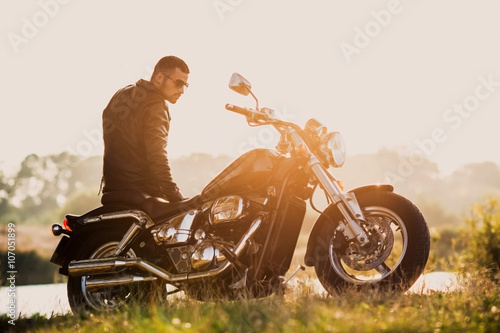 Canvas Print young brutal man in a black jacket and glasses standing near a motorcycle