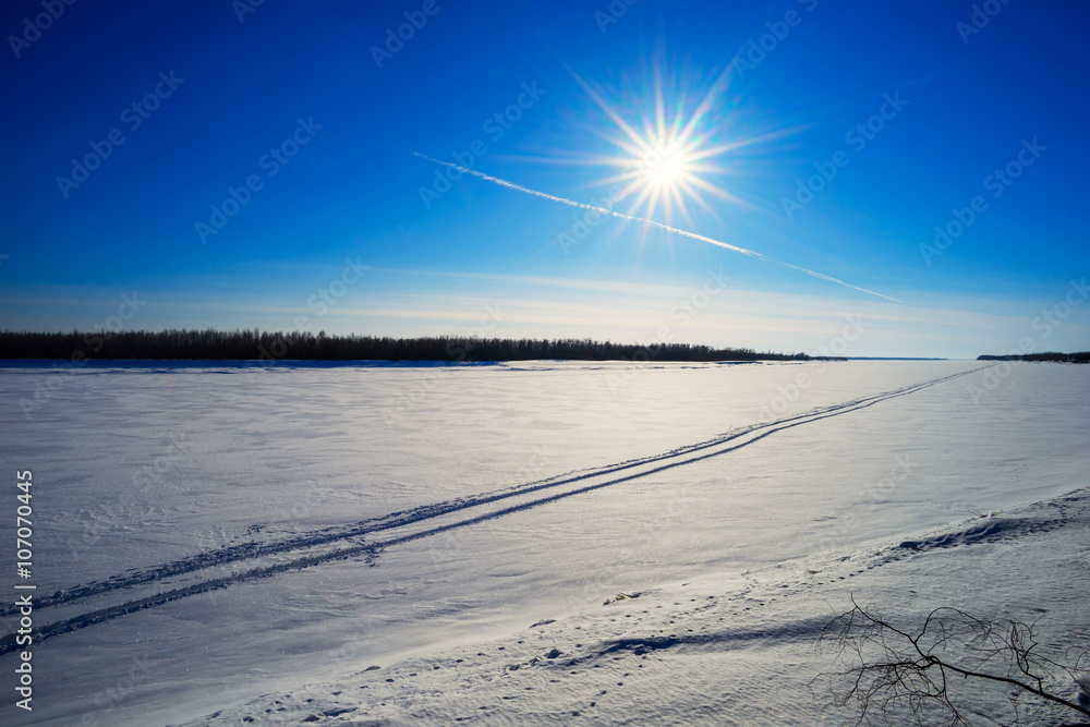 Traces of an airplane in the blue sky and from all-terrain vehicle on  frozen river .