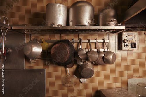 Kitchen utencils in old vintage professional bakery. Heavy used industrial pans, pots, whisks and more isolated on steel shelf and hangovers on wall with ceramic bricks