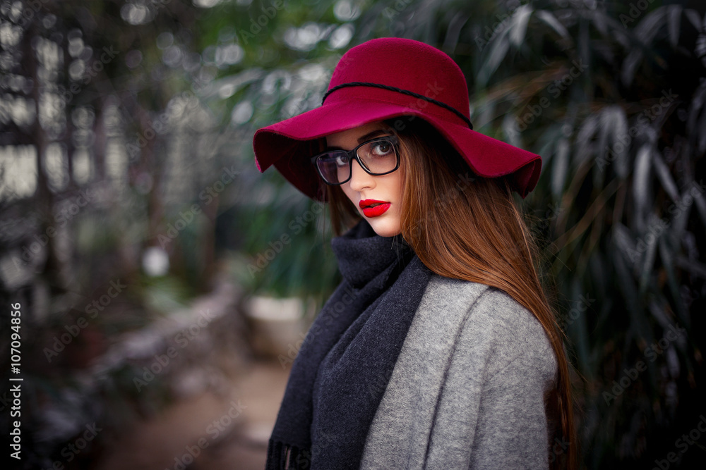 beautiful young brown-haired woman with long hair, beautiful makeup with bright red lips, wearing a gray coat, black pants, a scarf, a hat and sunglasses walking in a greenhouse
