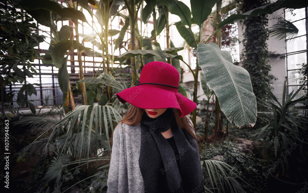 beautiful young brown-haired woman with long hair, beautiful makeup with bright red lips, wearing a gray coat, black pants, a scarf, a hat and sunglasses walking in a greenhouse