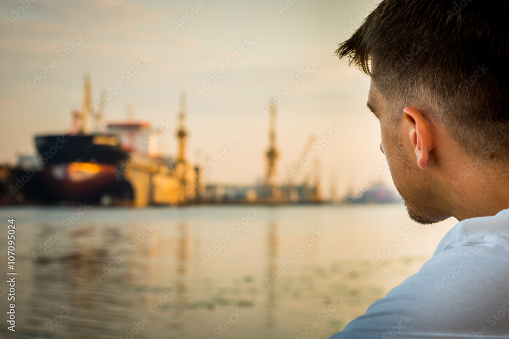 Fototapeta premium Head of the young man sitting on the bank of the river with a blurred ship in the background