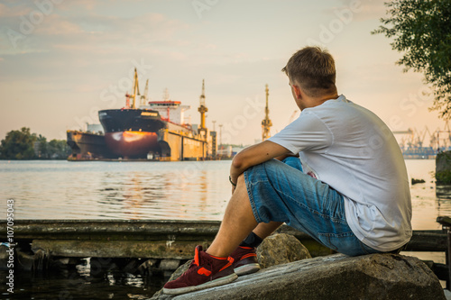 Young man sitting on the bank of the river with a blurred ship in the background, Szczecin, Poland photo