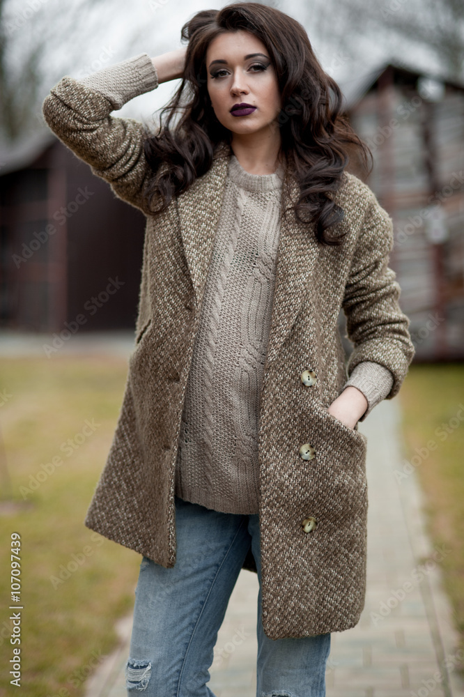 Spring in the country style. Full length portrait of young woman in white knitted sweater and furry hat standing in the front of rustic wood wall in the yard near house, wearing sweater casual. Spring in the country style. Full length portrait of young woman in white knitted sweater and furry hat standing in the front of rustic wood wall in the yard near house, wearing sweater casual.