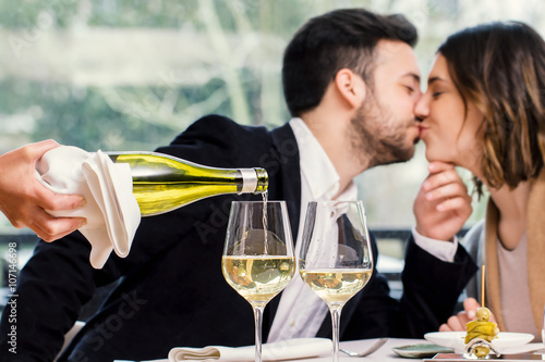Sommelier pouring wine with kissing couple in background.