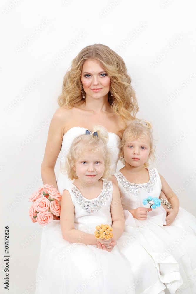 Beautiful blond mom with two young daughters blondes in white dresses on a white background. Beautiful mother and daughters in studio Beautiful blond mom with two young daughters blondes in white dresses on a white background. Beautiful mother and daughters in studio