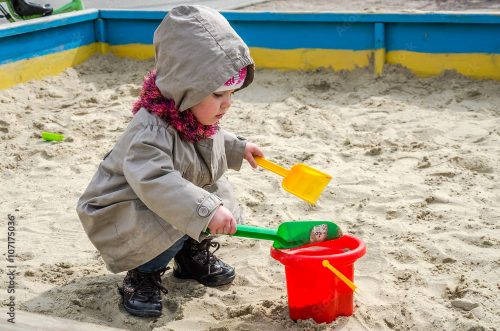 Little lovely girl baby playing in the sandbox on the playground with a shovel and bucket digging a hole, dressed in a raincoat with a hood, scarf and hat Little lovely girl baby playing in the sandbox on the playground with a shovel and bucket digging a hole, dressed in a raincoat with a hood, scarf and hat