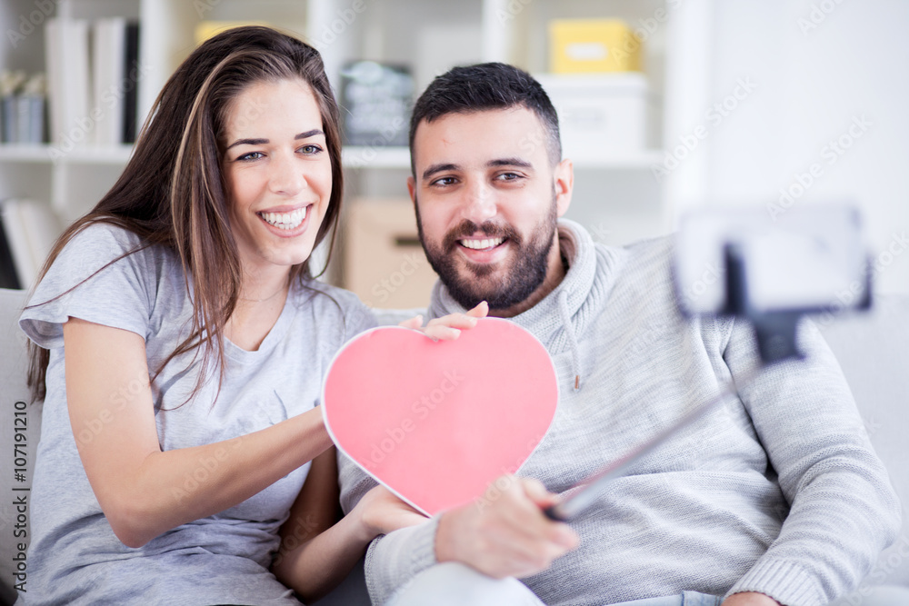 Happy young beautiful couple taking photo with smart phone on selfie stick. Young couple having fun in the living room