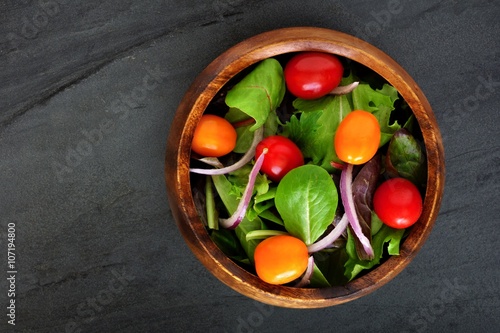 Garden salad with cherry tomatoes and red onions in wooden bowl overhead view on dark slate background
