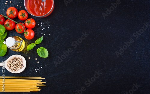 Pasta ingredients - tomatoes, olive oil, garlic, italian herbs, fresh basil, salt and spaghetti on a black stone background with copy space, horizontal, top view