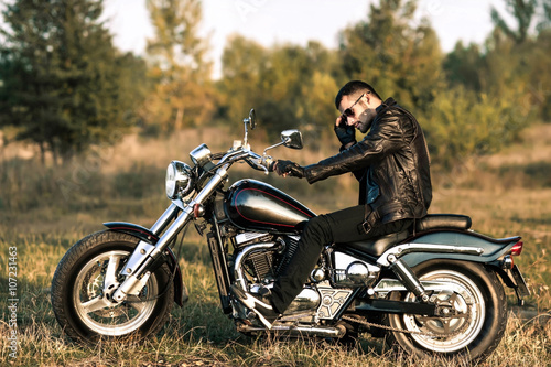 Fotografie young brutal man in a black jacket and glasses sits near a motorcycle