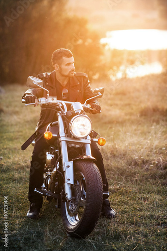 Fotografie young brutal man in a black jacket and glasses sits near a motorcycle