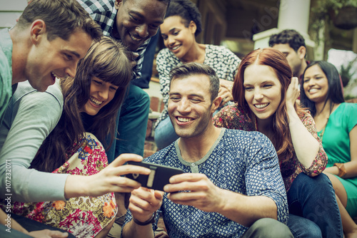 A group of friends on the steps of a house porch, looking at a smart phone selfy on the screen.