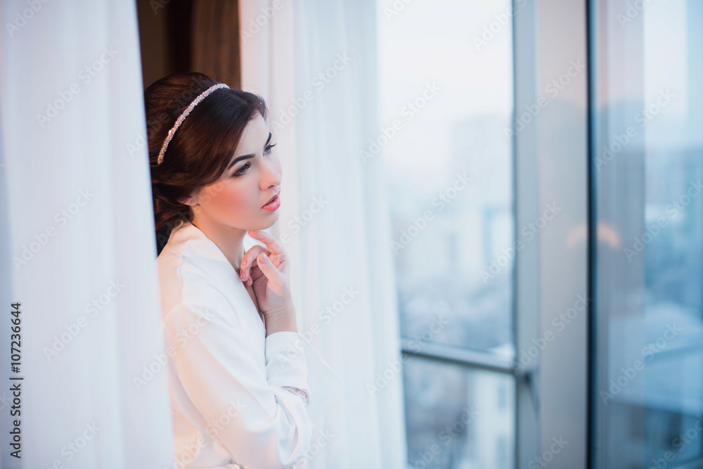 Portrait of a beautiful young girl in a white robe in the bedroom near the window, morning, boudoir, relaxation, evening