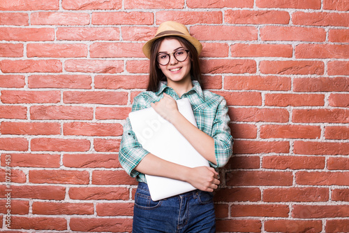 Beautiful young dark-haired girl in casual clothes, hat and eyeglasses posing, smiling and with close laptop in hands, standing against brick wall