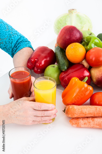 Hands of mature female putting two glasses with red and yellow juice on the table near vegetables and fruits