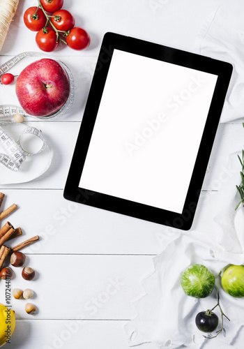 Tablet with a measuring tape, rosemary, lime, lemon, nuts, tomatoes, cinnamonnand red apples on a white wooden background top view vertical photo