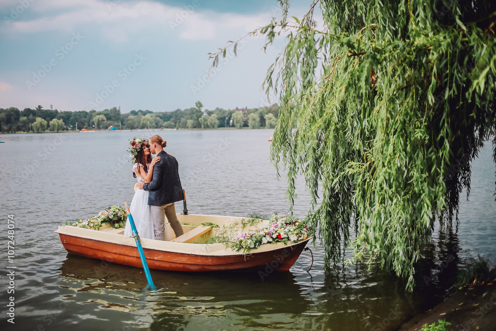 bride and groom sitting in an orange row boat floating out of the tree covering and in to the lake. Bride with wreath of flowers. Couple hugging and kissing