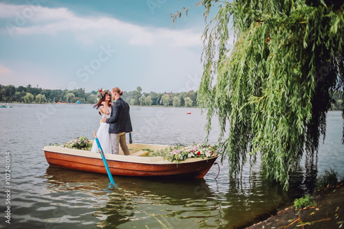 bride and groom sitting in an orange row boat floating out of the tree covering and in to the lake. Bride with wreath of flowers. Couple hugging and kissing © nataliakabliuk