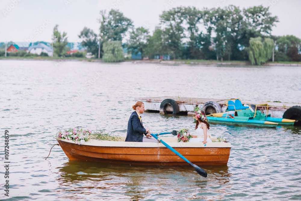 stylish gorgeous happy brunette bride and elegant groom in the boat on the background of a lake. Bride with wreath of flowers stylish gorgeous happy brunette bride and elegant groom in the boat on the background of a lake. Bride with wreath of flowers
