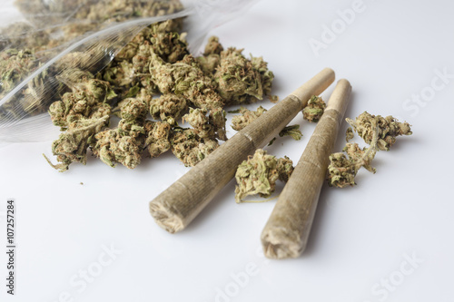 Pile of medical cannabis dried buds scattered from nylon package and two marijuana joints on white background from side