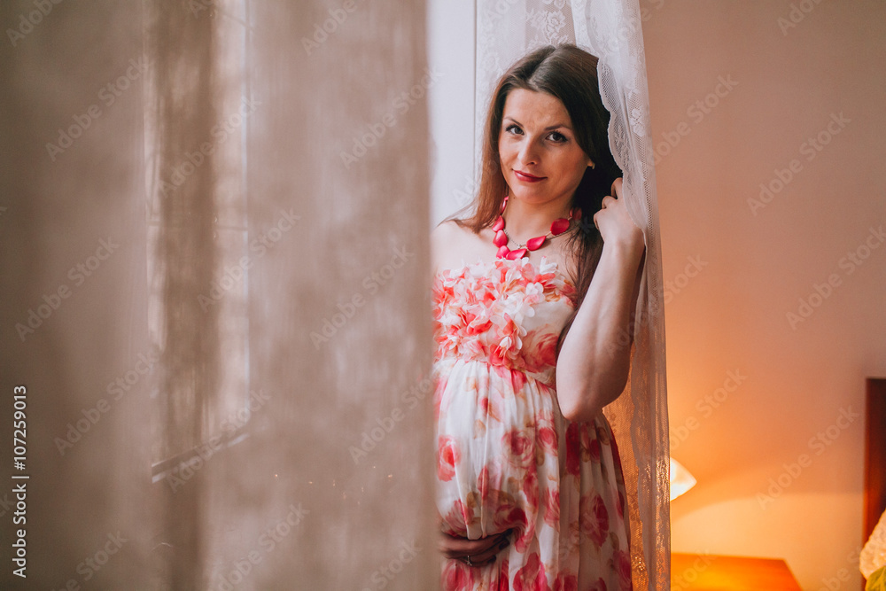 young pregnant woman staying near the window in pink dress