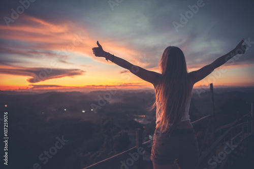 Obraz na plátně Silhouette of the woman spreading arms with her thumbs up, standing high on the viewpoint with breathtaking view over fields in sunset light